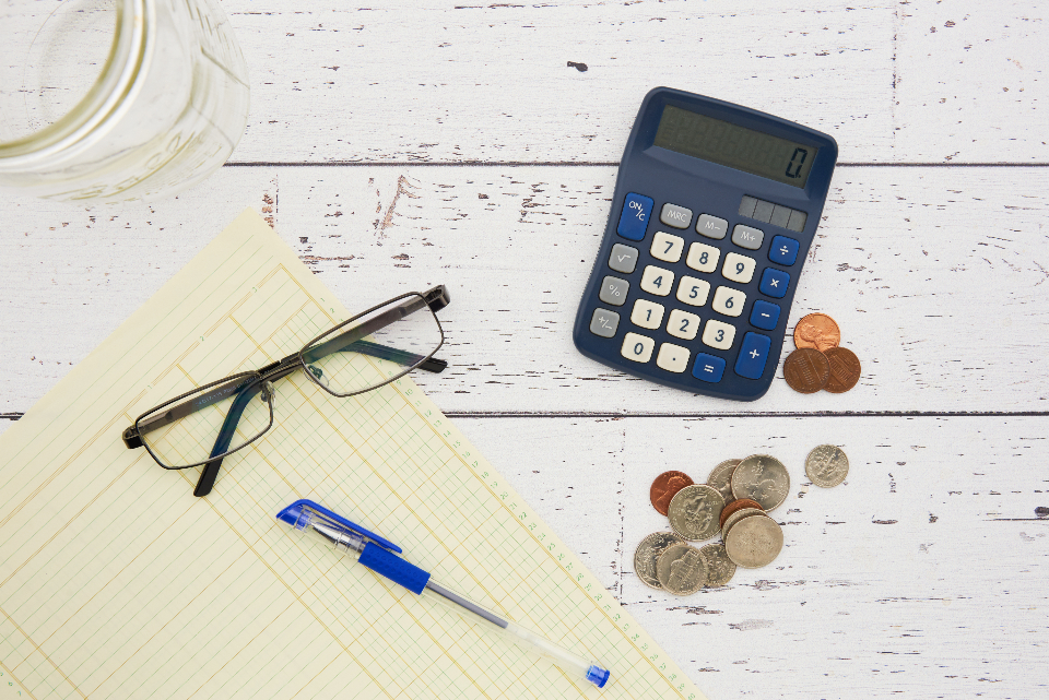 A calculator, reading glasses, a pen and a few coins on a ledger pad